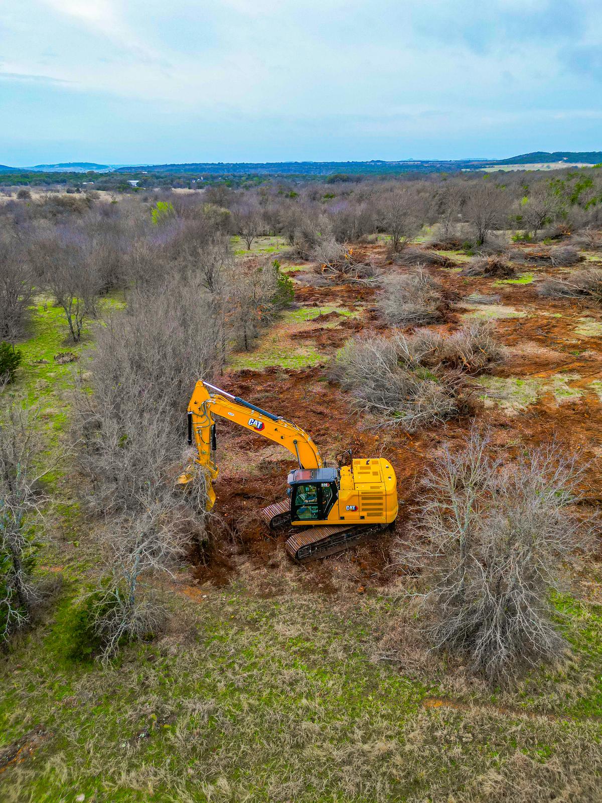 Aerial view of a tracked excavator clearing a lane through heavy brush, with windrowed debris and turned red dirt behind it