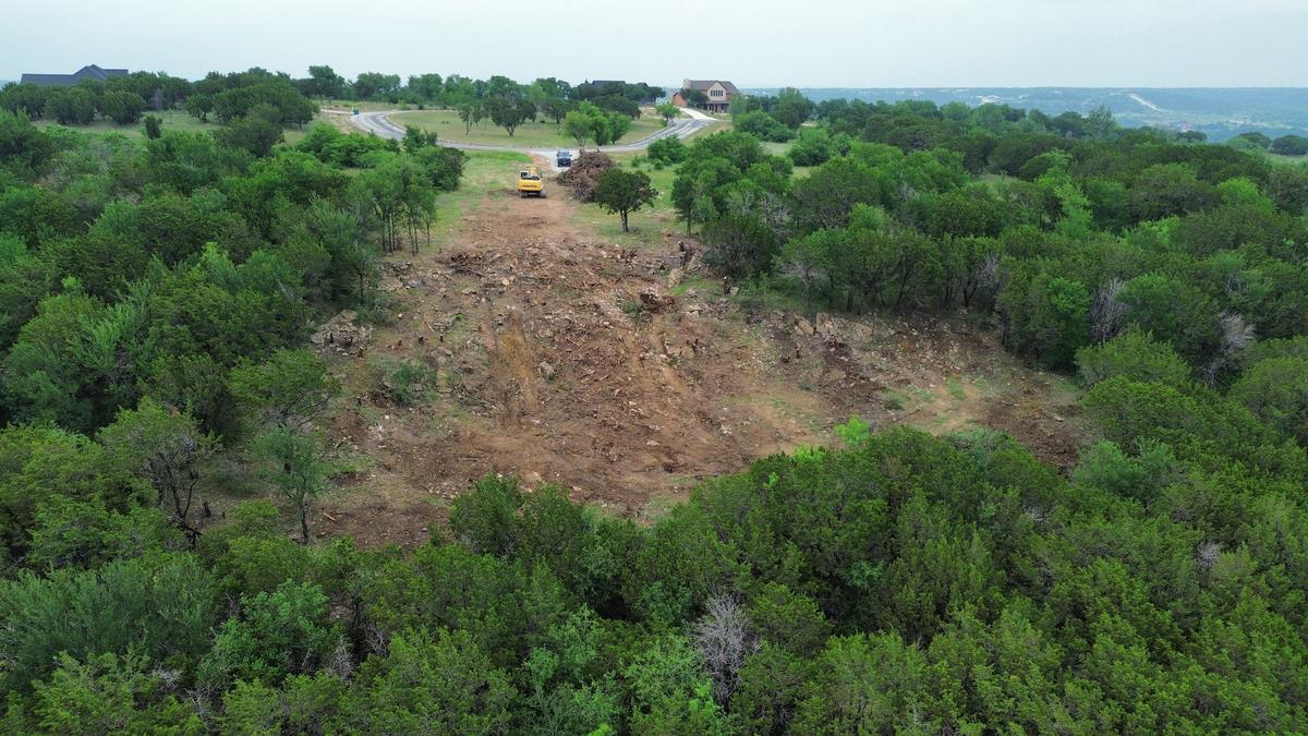 Aerial view of a cleared swath cut through dense cedar on a ridge above Possum Kingdom Lake, with an excavator and a brush pile at the road end