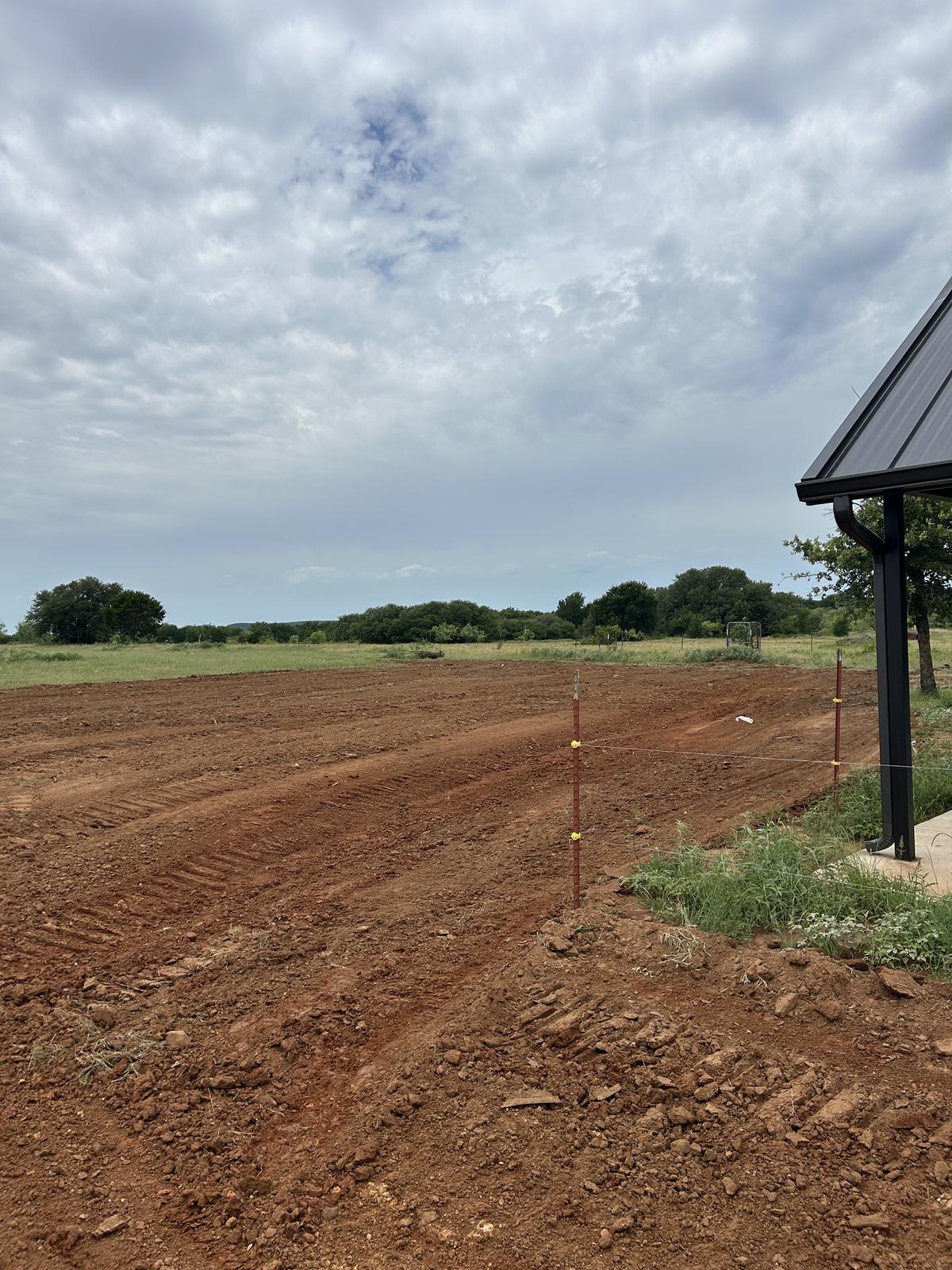 A large area of freshly cleared and graded red dirt beside a new metal building near Graham, Texas, with open pasture and a tree line beyond