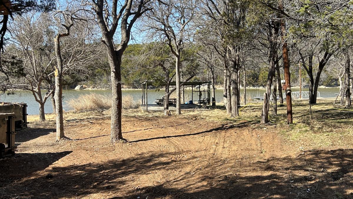 The same lakeside lot cleared to bare, leveled dirt with the mature trees left standing and a boat dock visible on the water beyond