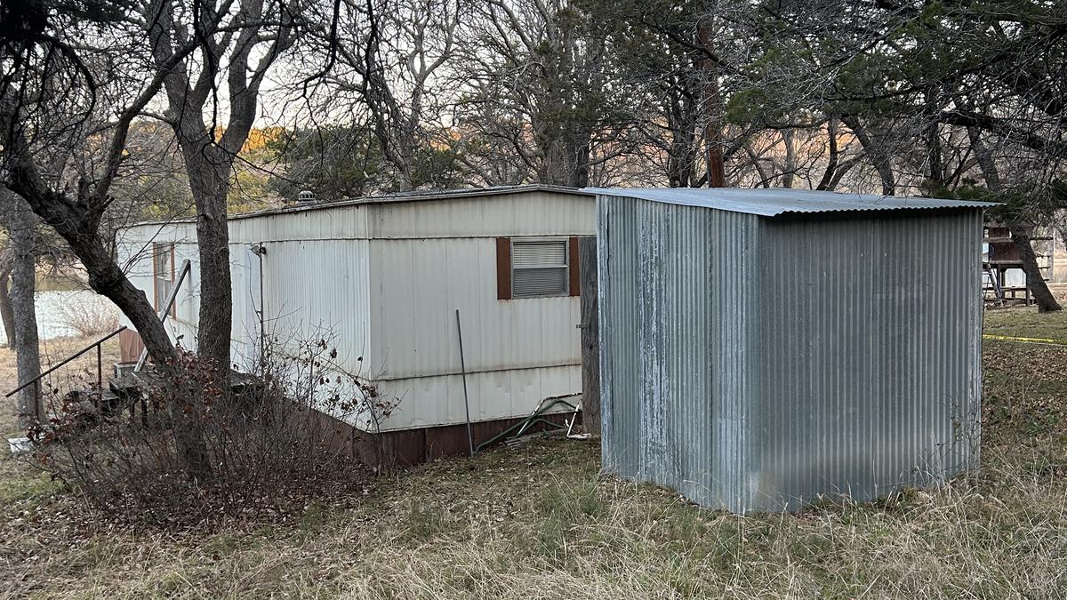 An old white mobile home and a corrugated metal shed standing in overgrown brush among bare winter trees near a lake