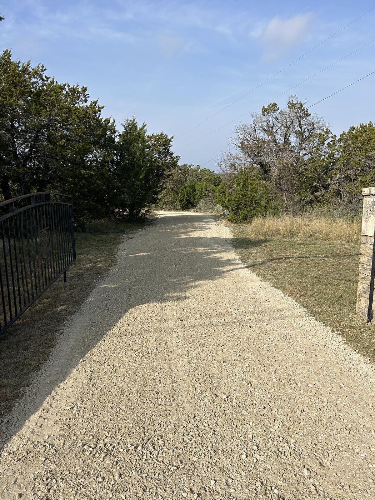 A newly laid crushed limestone driveway running through an iron gate and stone column, climbing through cedar toward the top of a rise
