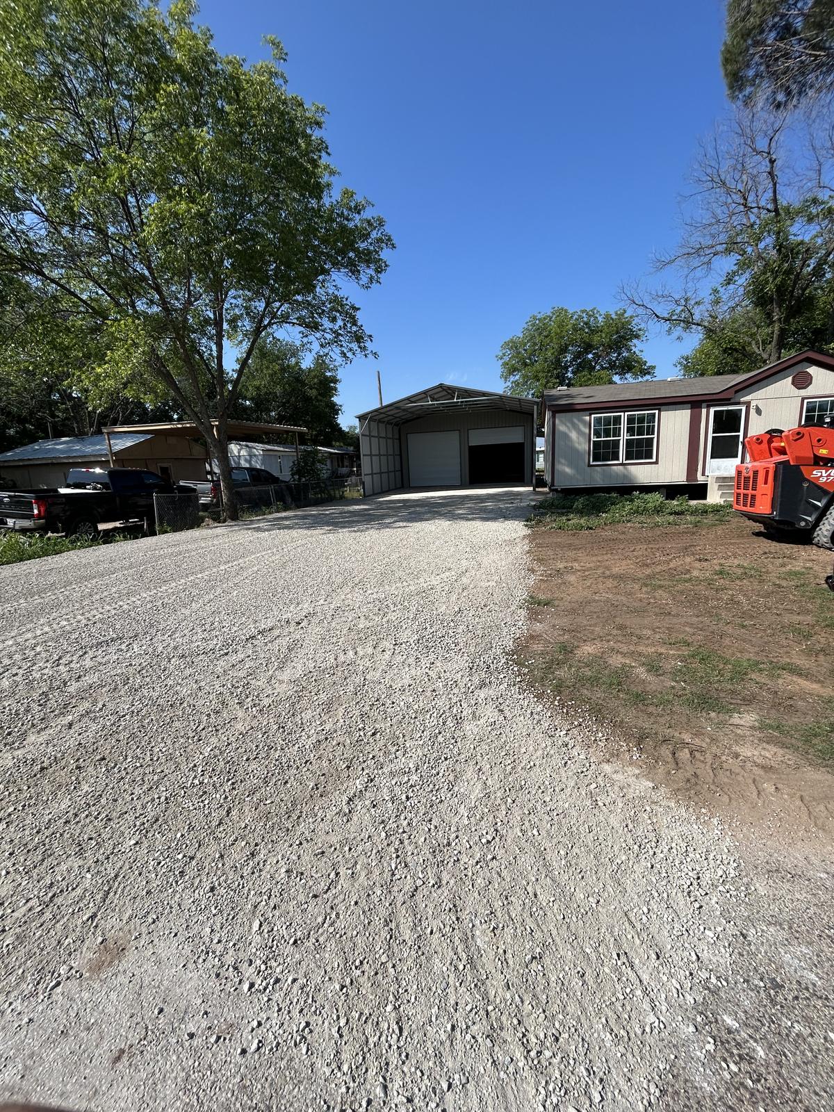 A freshly rocked gravel driveway in Graford, Texas running up to an open metal shop beside a home, with a track loader parked at the edge