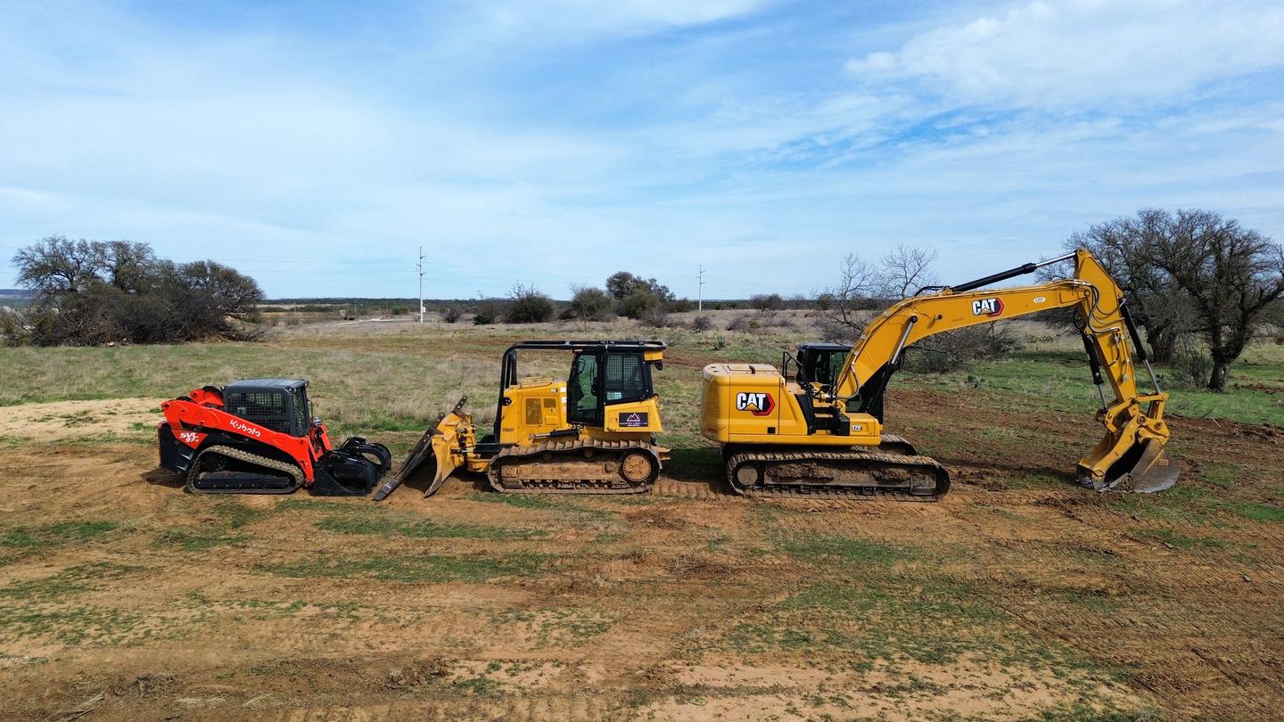 A Kubota track loader, a Cat dozer and a Cat 320 excavator lined up side by side in a pasture