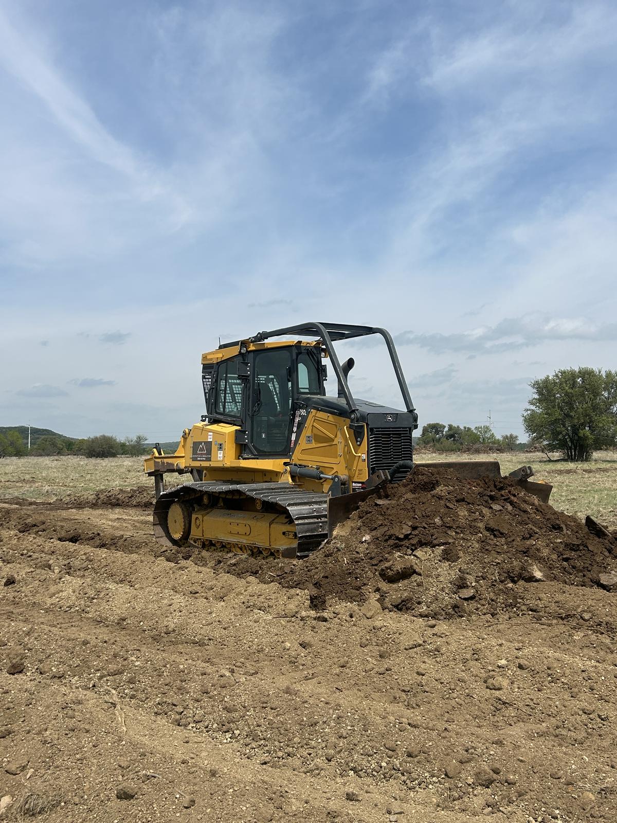A John Deere 750L dozer pushing a windrow of dark dirt while building up a building pad in a pasture