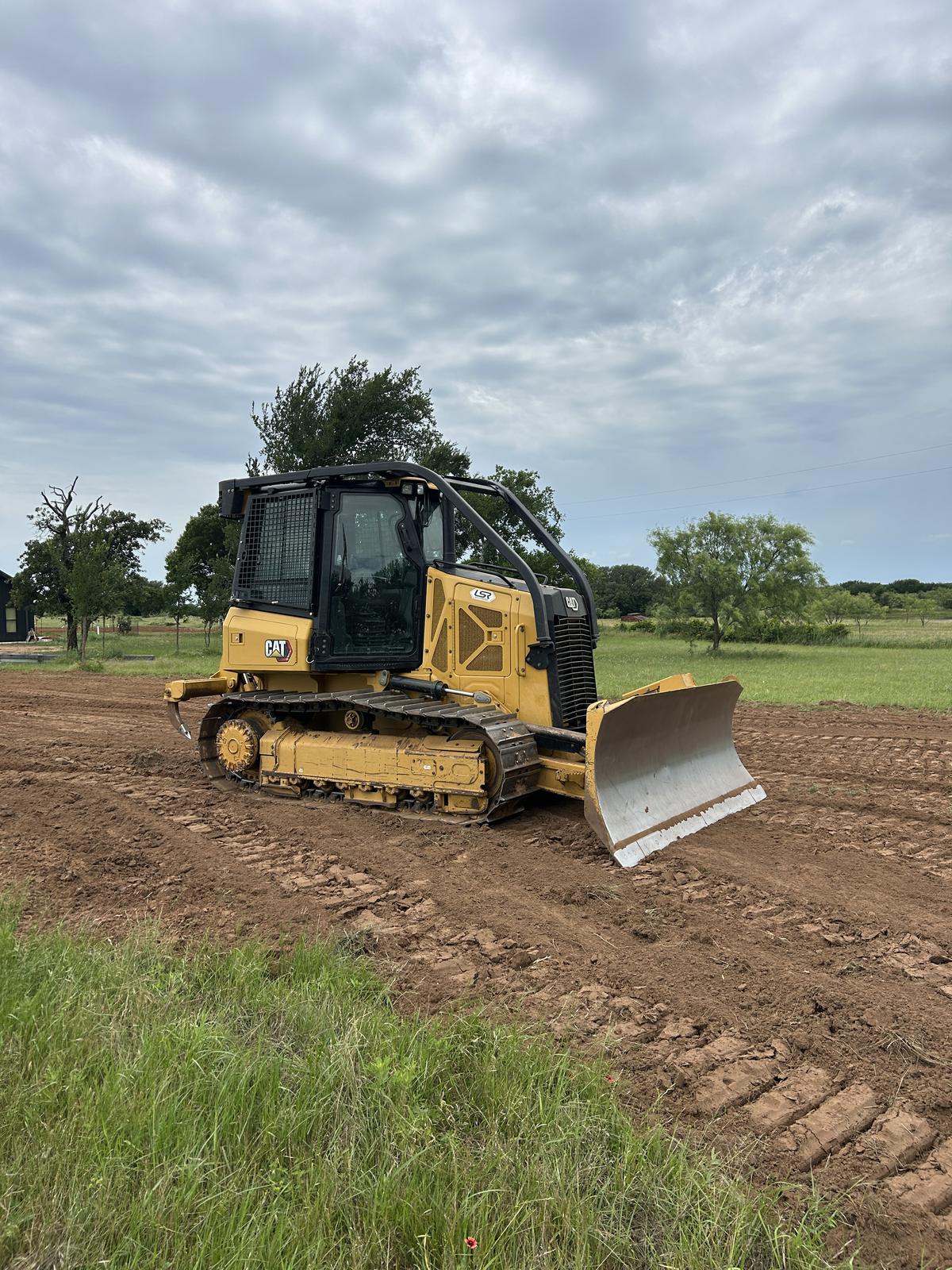 A Cat dozer with its blade down on freshly worked brown dirt beside a building site, under an overcast sky