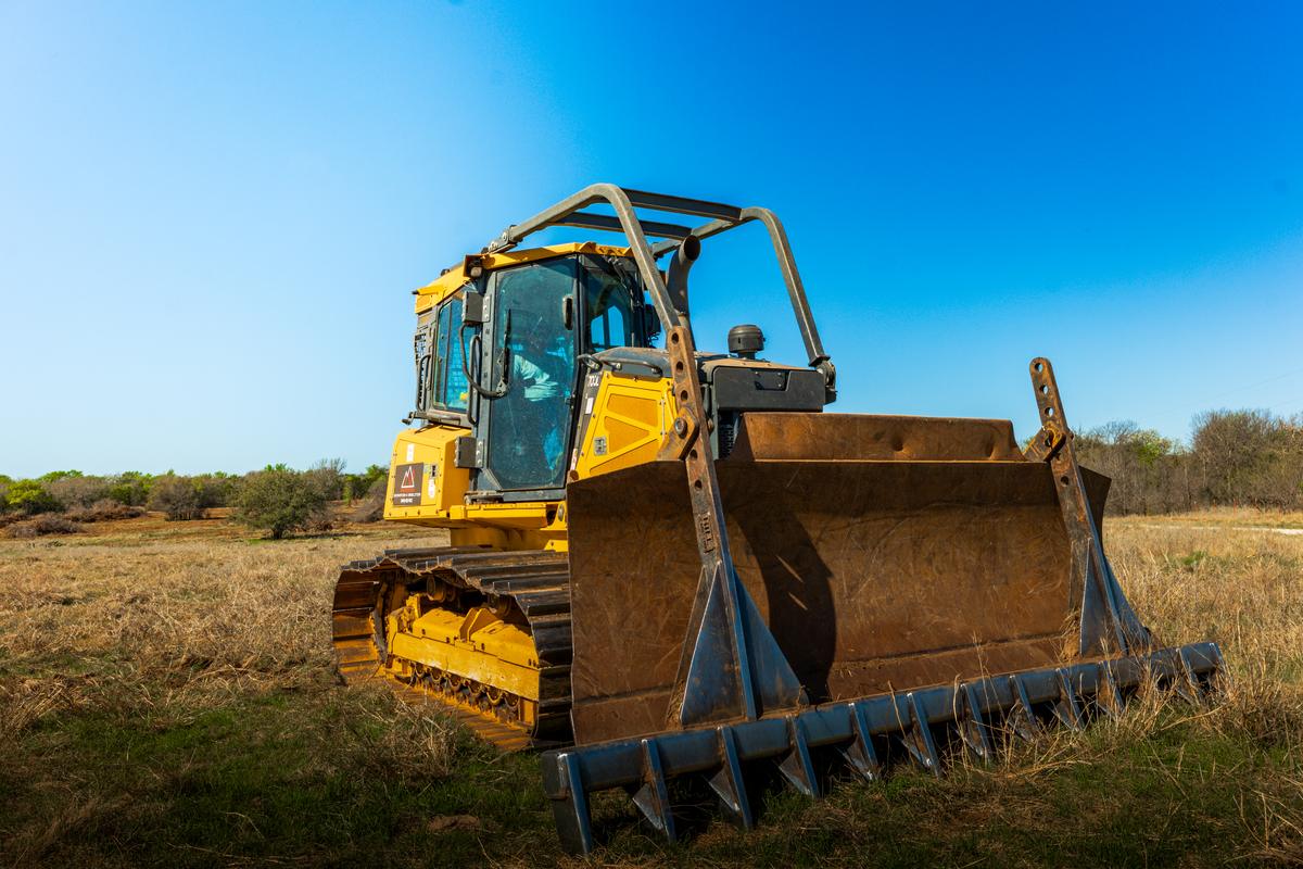 A Cat dozer fitted with a wide root rake blade, parked in dry grass under a clear blue sky