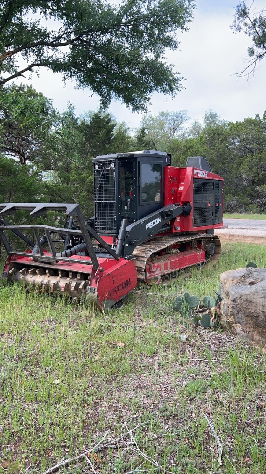 A red Fecon tracked forestry mulcher parked in cedar country beside a gravel road, its drum head lowered into the grass