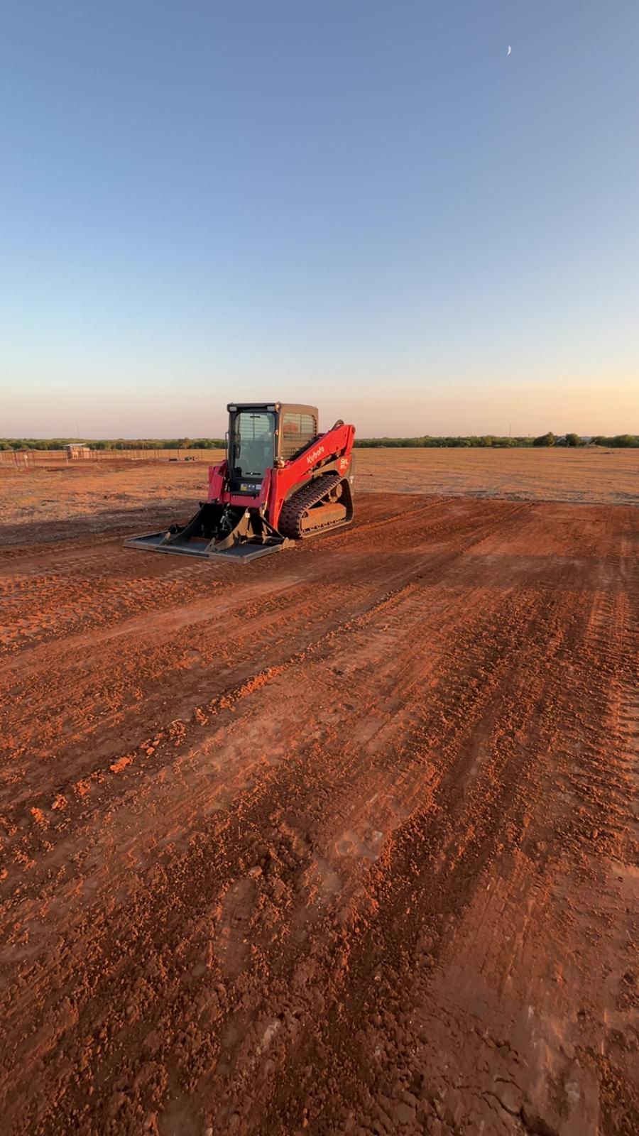 A red Kubota compact track loader with a land plane on a freshly graded red dirt building pad at sunset, with a crescent moon overhead