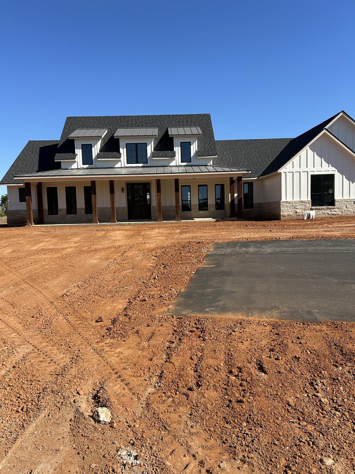 A newly built white farmhouse with a covered porch, standing on a graded red dirt site beside a fresh concrete driveway