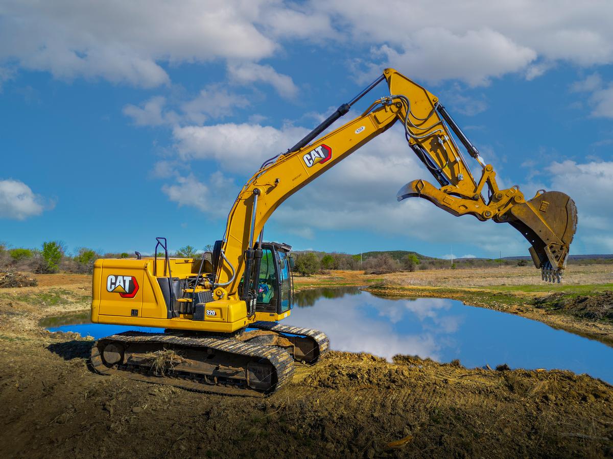 A Cat 320 excavator parked on the bank of a newly dug stock tank, the still water reflecting a blue sky and clouds