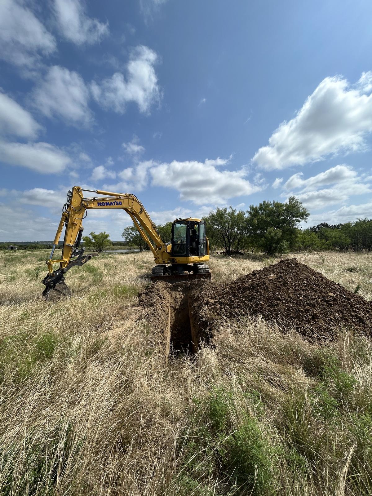 A Komatsu excavator digging a narrow, straight trench across dry pasture grass with the excavated dirt windrowed alongside and a stock tank beyond