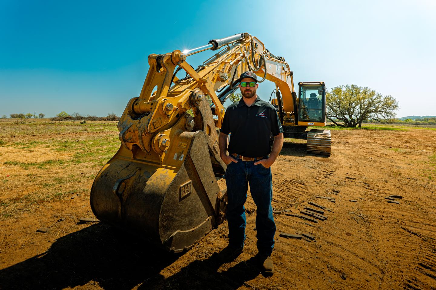 Trey Ranft standing in front of the bucket of his Cat excavator on a worked dirt site